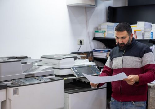 a man is standing behind printer copiers with a printer paper on his hands